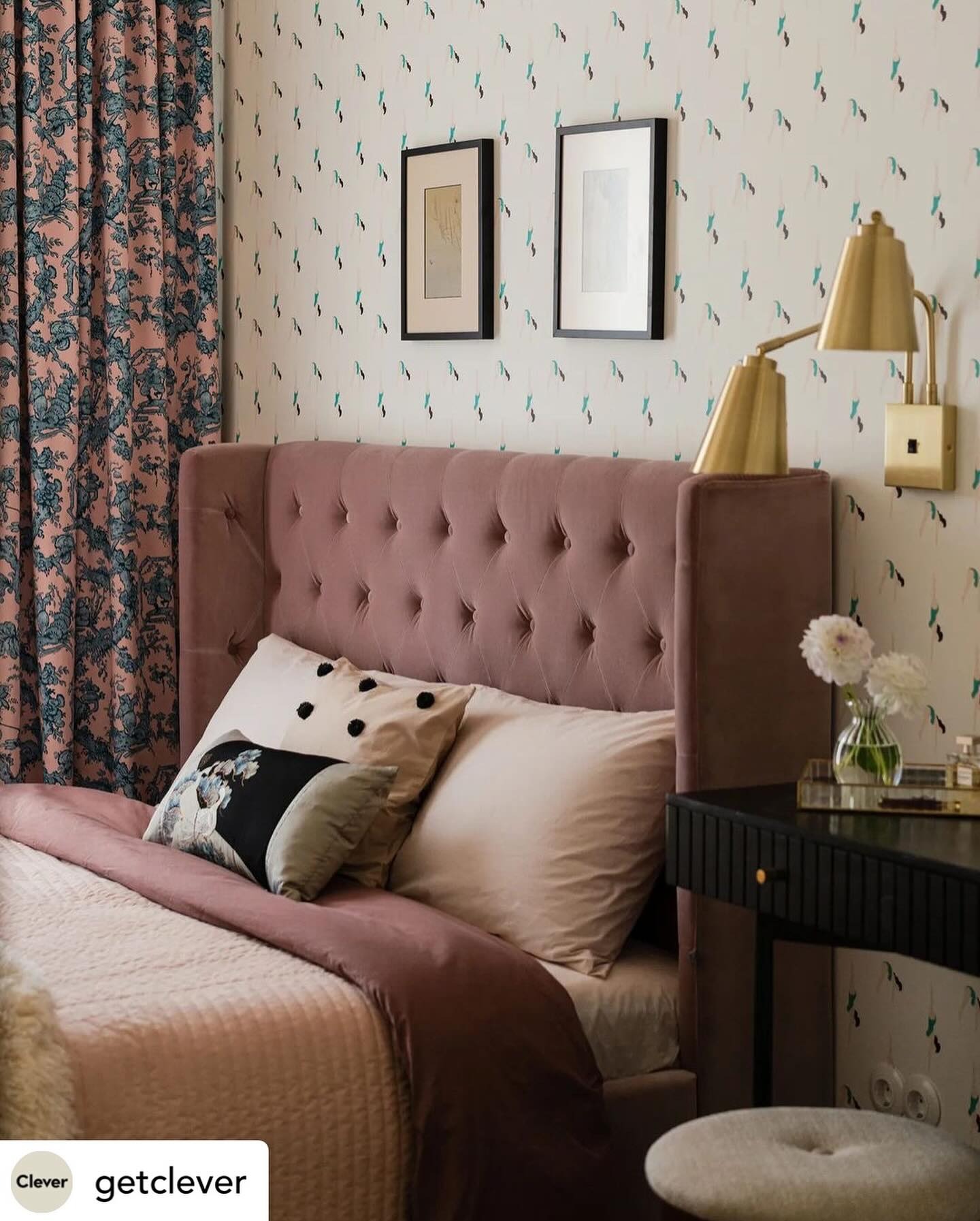 Różowe łóżko z wezgłowiem w stylu Chesterfield, obok czarna toaletka z bukietem białych kwiatów, na ścianie tapeta w drobne wzory i złota lampka.