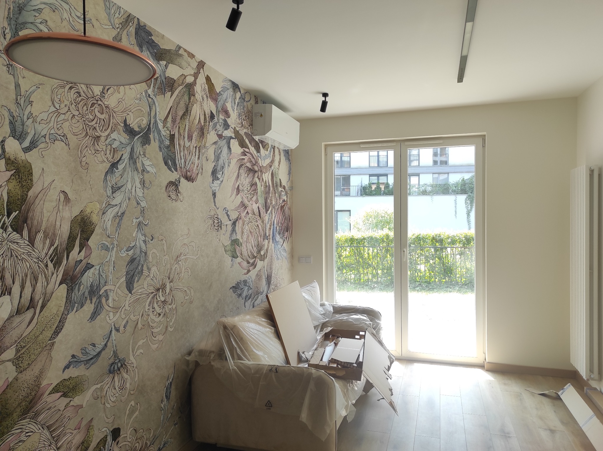 Pokój po remoncie z ozdobną tapetą w kwiaty, nowym oświetleniem sufitowym, klimatyzacją i kanapą zabezpieczoną folią ochronną oraz elementami zabudowy meblowej do montażu.