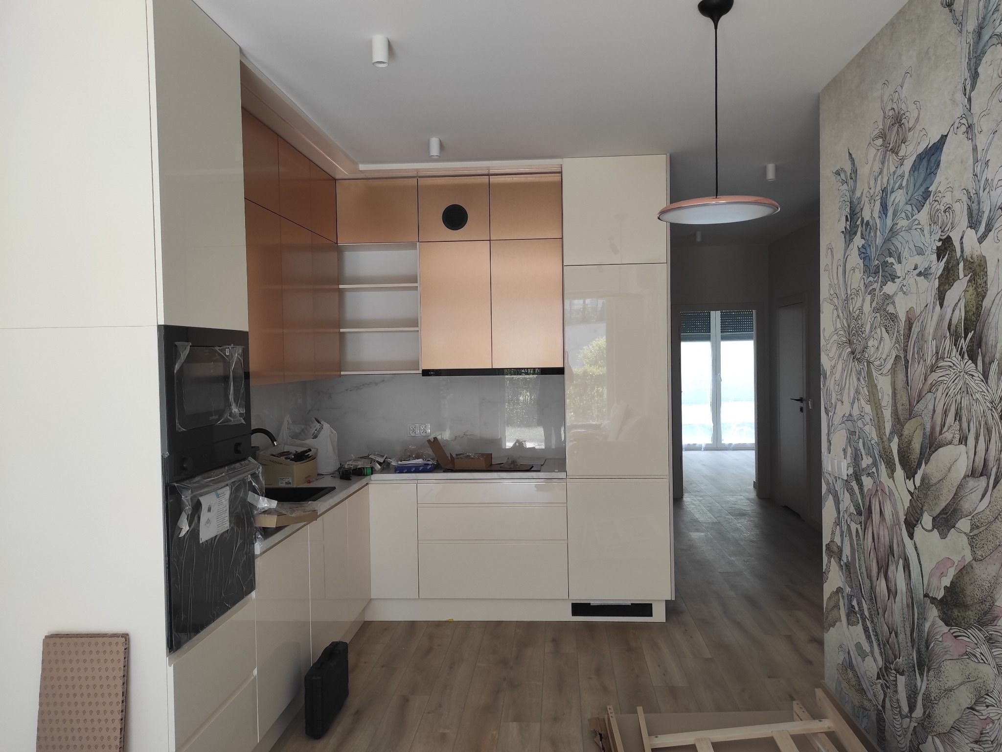 Nowoczesna kuchnia w trakcie montażu: kremowe szafki z elementami w odcieniach miedzi, sprzęt AGD w folii ochronnej, kartony na blacie, podłoga w panelach, ściana z tapetą w kwiatowe wzory.