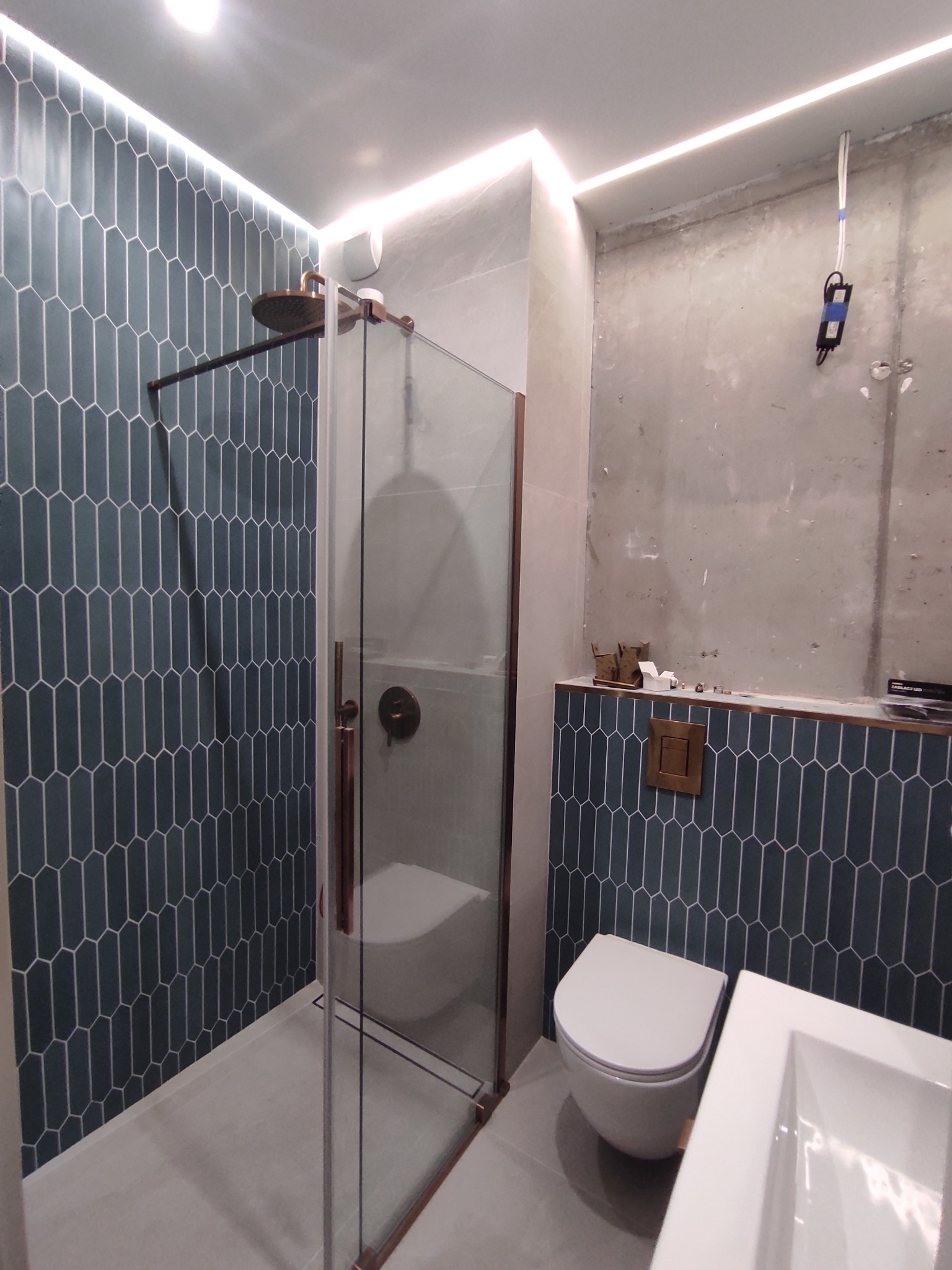 Nowoczesna łazienka w trakcie wykańczania: kabina prysznicowa z miedzianymi detalami, toaleta podwieszana i umywalka na tle niebieskich płytek w kształcie wydłużonego sześciokąta oraz surowej...