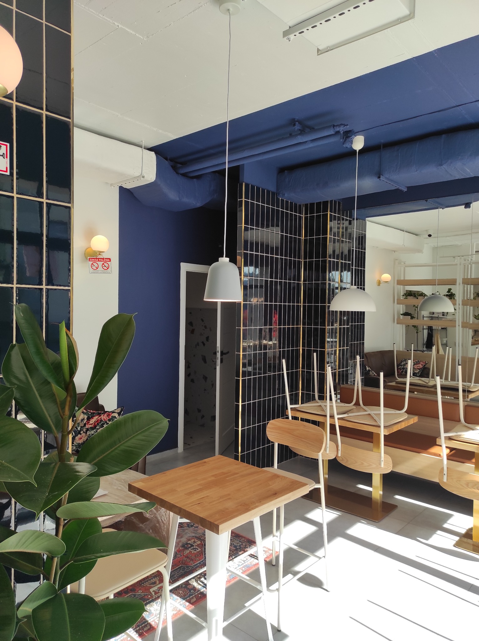 Wnętrze kawiarni z niebieskim sufitem, czarnymi płytkami z złotymi fugami, drewnianymi stolikami i krzesłami odwróconymi na blatach, z rośliną doniczkową w lewym dolnym rogu i znakiem zakazu...