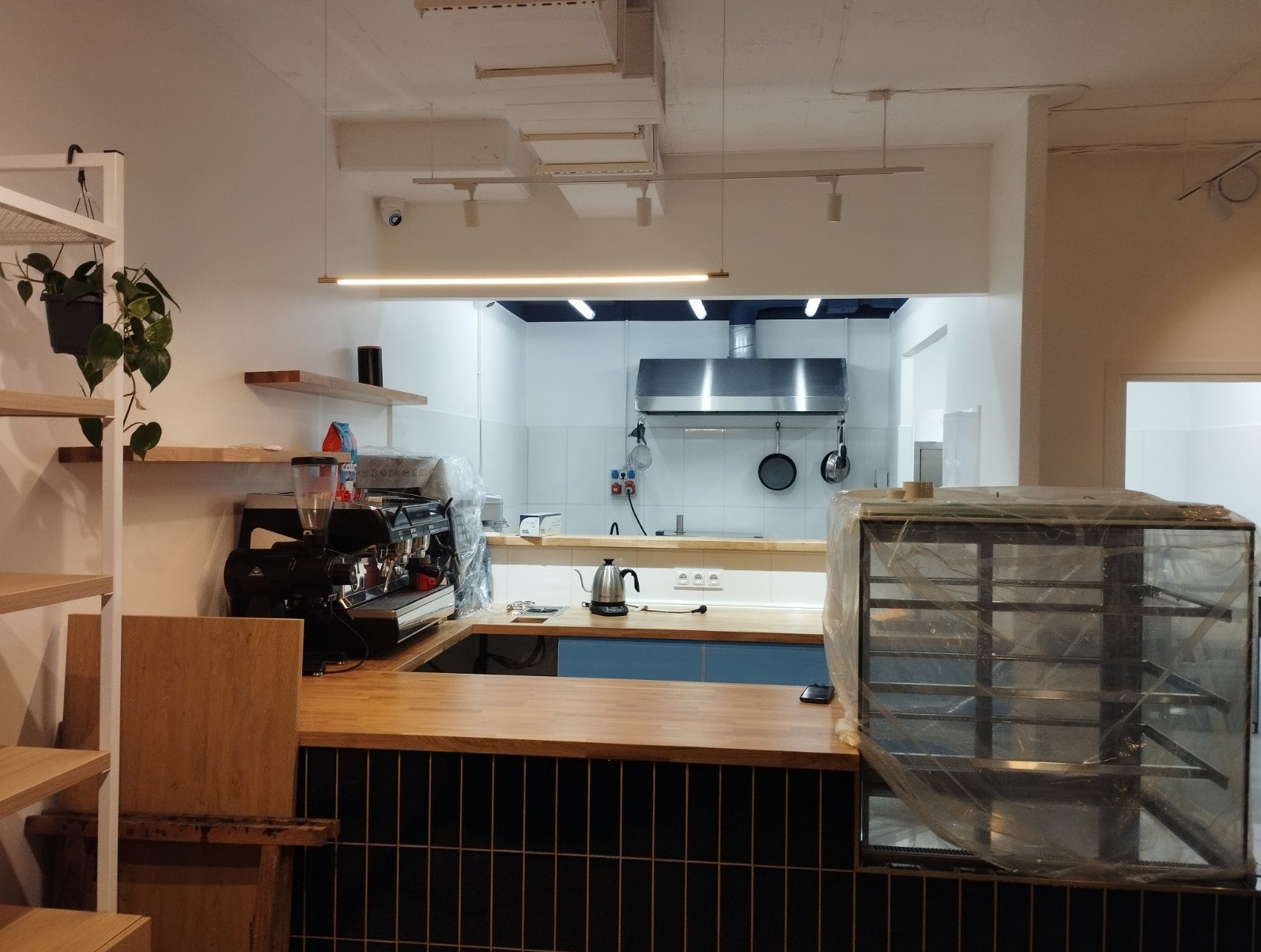 Wnętrze lokalu gastronomicznego w trakcie wykańczania: widoczny ekspres do kawy, lada z czarnymi płytkami, stalowa kuchnia z okapem i wiszącymi patelniami, półki z roślinami i przykryta folią...