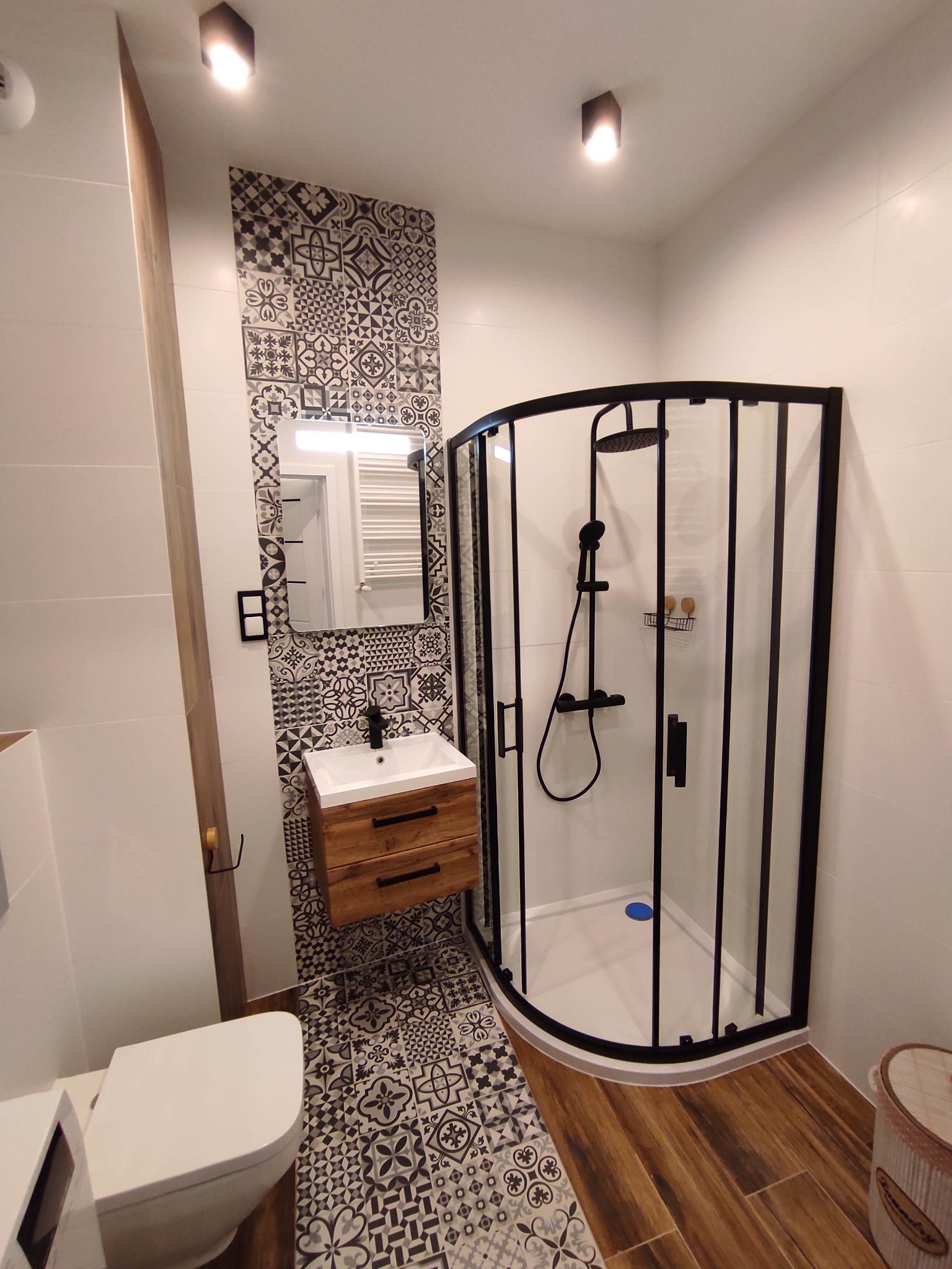 Nowoczesna łazienka z kabiną prysznicową w czarnej ramie, drewnianą szafką pod umywalką i mozaiką na ścianie oraz podłodze. Widoczne białe płytki na ścianach i drewnopodobne panele podłogowe.