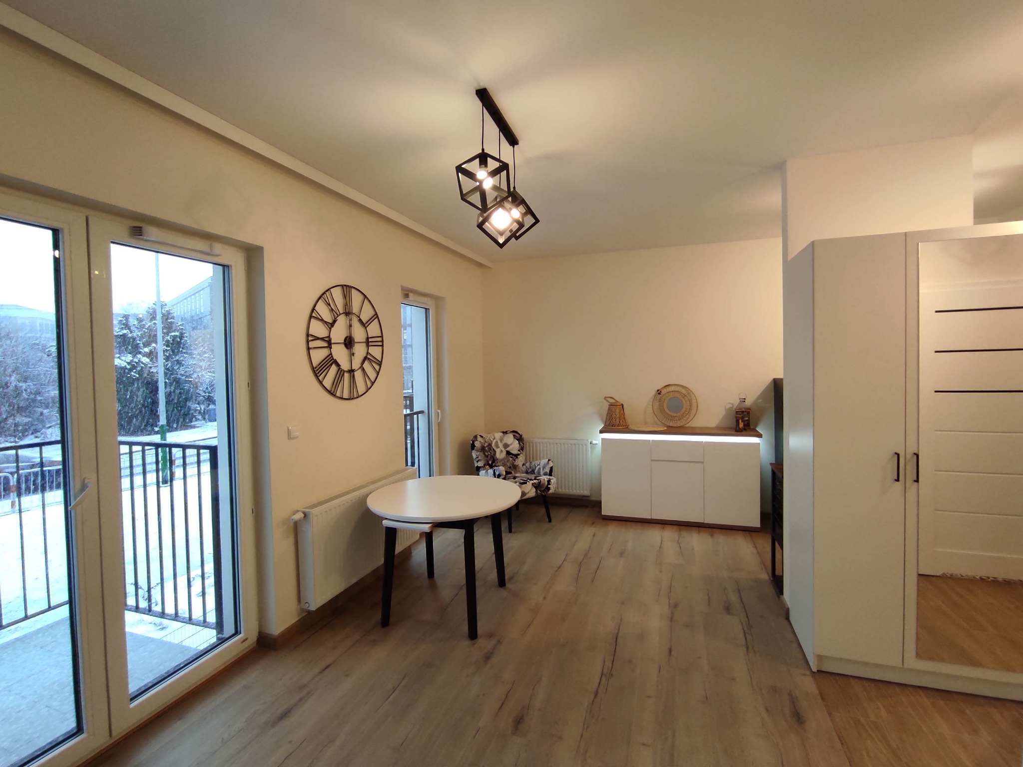 Jasne wnętrze mieszkania z drewnianą podłogą, okrągłym stołem z czarnymi nogami, nowoczesną lampą sufitową z geometrycznymi kloszami, dużym zegarem na ścianie i widokiem na zaśnieżony balkon