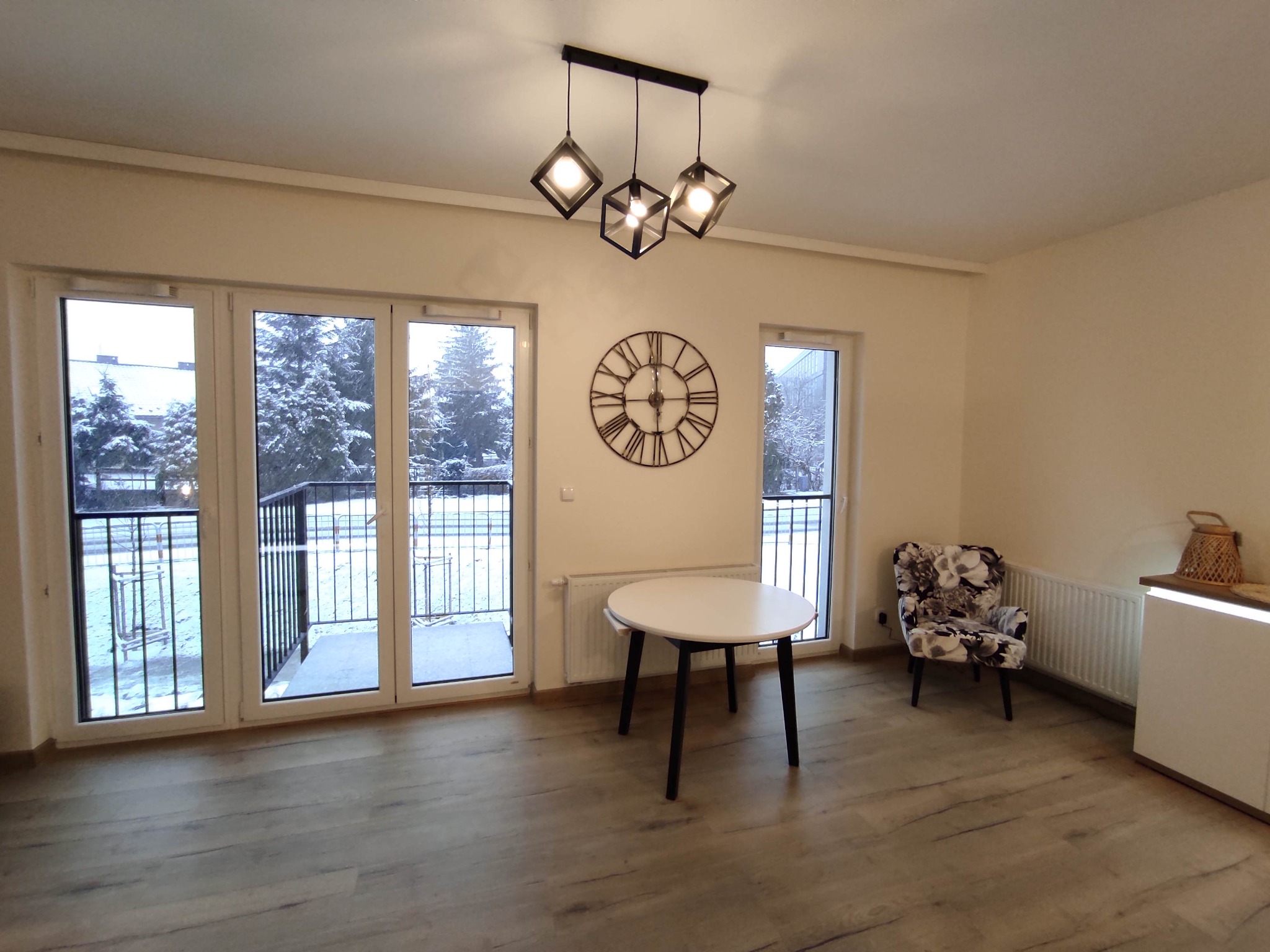 Jasne wnętrze z drewnianą podłogą, okrągłym stołem na czarnych nogach, fotel z motywem kwiatowym i balkonem z widokiem na zaśnieżone drzewa