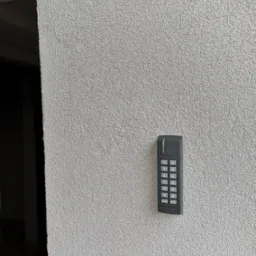 Szary panel numeryczny domofonu zamontowany na teksturowanej ścianie w pobliżu wejścia.