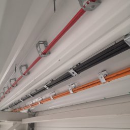 PRO-Elektro Serhii Kopylov - Różnokolorowe kable (czerwony, czarny, pomarańczowy) przymocowane metalowymi uchwytami do białego sufitu z blachy trapezowej w hali przemysłowej.