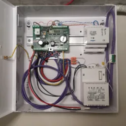 Otwarta skrzynka z elektroniką zabezpieczeń: płyta główna, transformator Pulsar AWT150, moduł komunikacyjny Roger UT-4DR oraz liczne przewody w kolorach fioletowym, białym, czarnym i czerwonym.