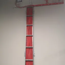 Pionowy kanał kablowy z dużą ilością czerwonych przewodów, zamontowany na jasnej ścianie, z widocznymi puszkami elektrycznymi na górze.