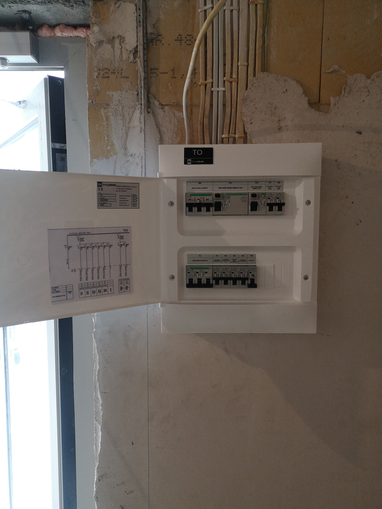 Otwarta skrzynka bezpiecznikowa zamontowana na surowej ścianie, widoczne przewody elektryczne i schemat instalacji na wewnętrznej stronie drzwiczek.