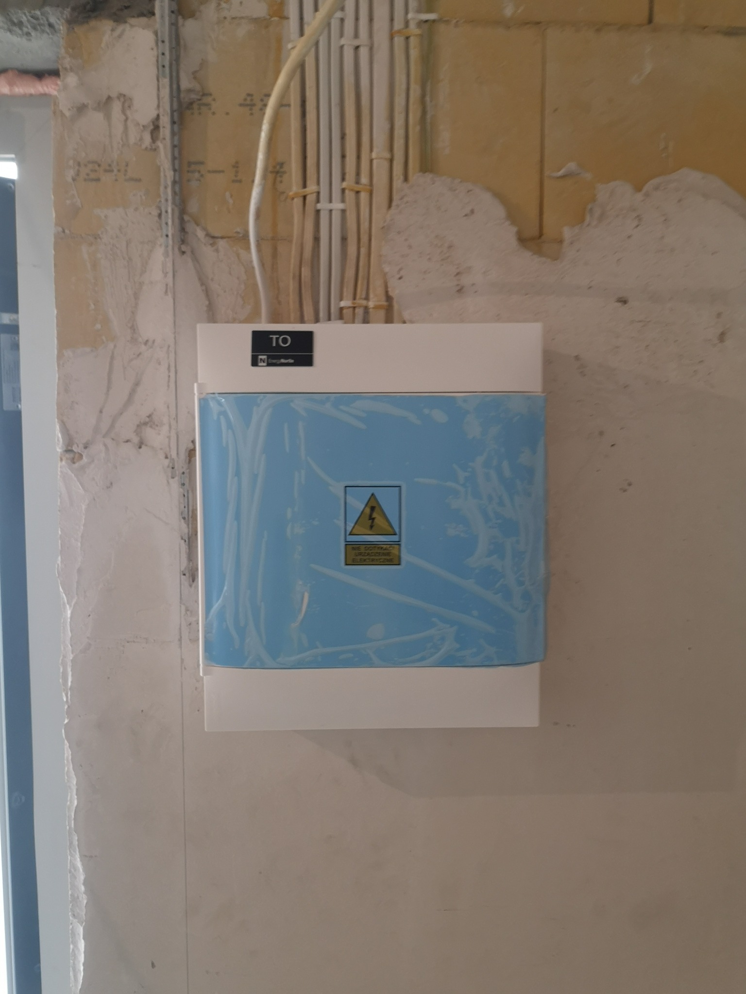Skrzynka elektryczna zabezpieczona folią ochronną, zamontowana na surowej ścianie w trakcie remontu, widoczne kable w peszlach.