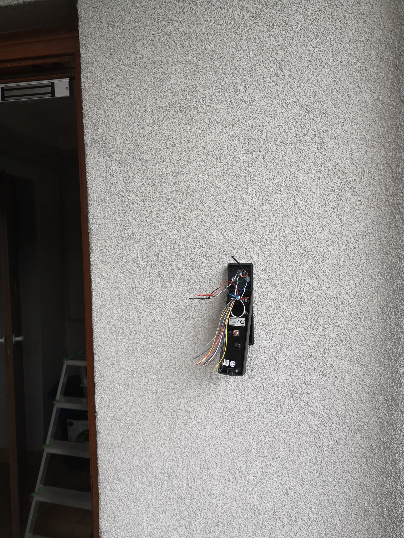 Montaż domofonu na elewacji budynku z widocznymi przewodami i elementami elektroniki, drabina w tle.