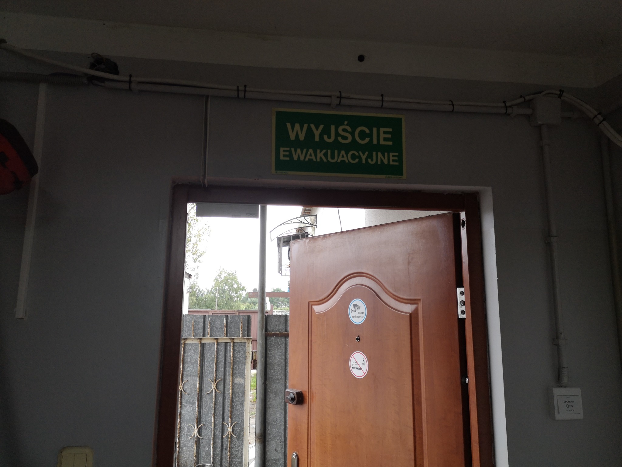 Otwarta brązowa brama ewakuacyjna z naklejkami informacyjnymi 'Obiekt monitorowany' i zakaz palenia, widoczna metalowa krata i zielony znak 'Wyjście Ewakuacyjne' nad drzwiami.