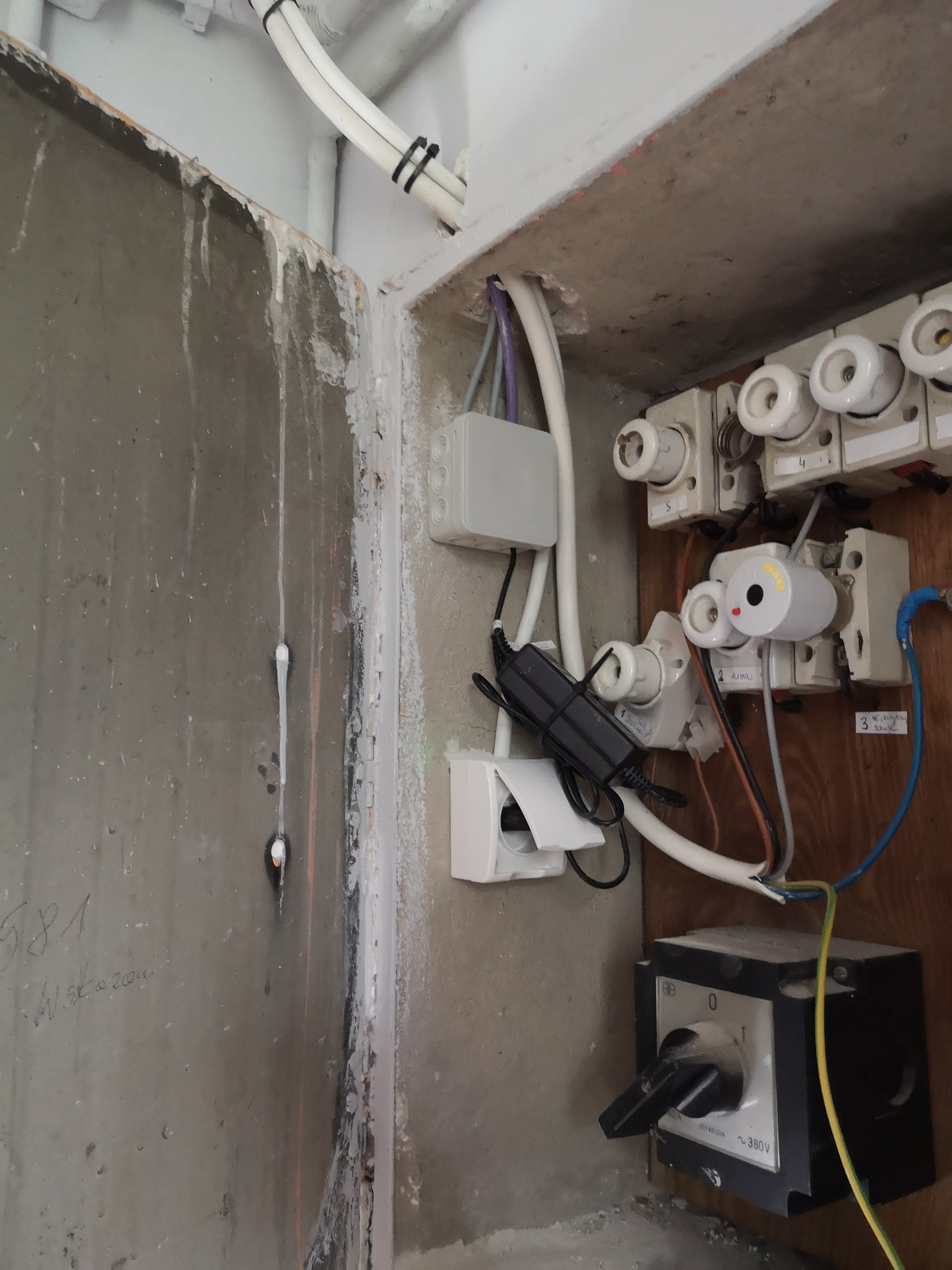 Otwarta skrzynka elektryczna z bezpiecznikami topikowymi, wyłącznikiem i gniazdkiem, widoczne przewody i puszki instalacyjne, betonowa ściana z zaciekami.