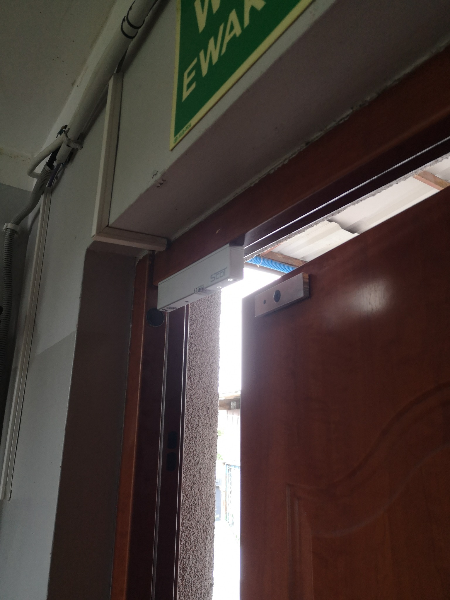 Elektromagnetyczny zamek do drzwi zamontowany nad brązowymi drzwiami, widoczna instalacja elektryczna i niebieski peszel.