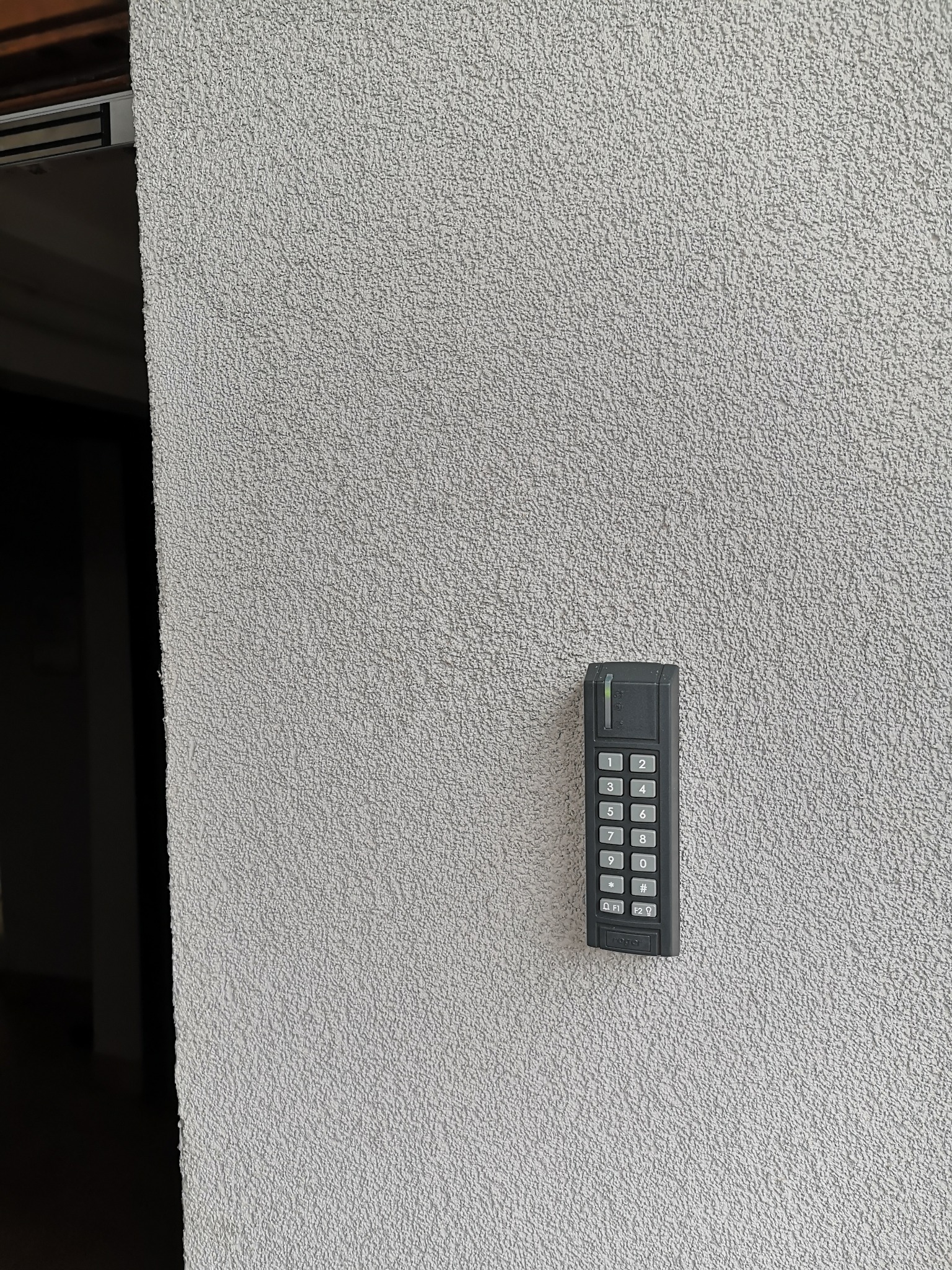 Szary panel numeryczny domofonu zamontowany na teksturowanej ścianie w pobliżu wejścia.