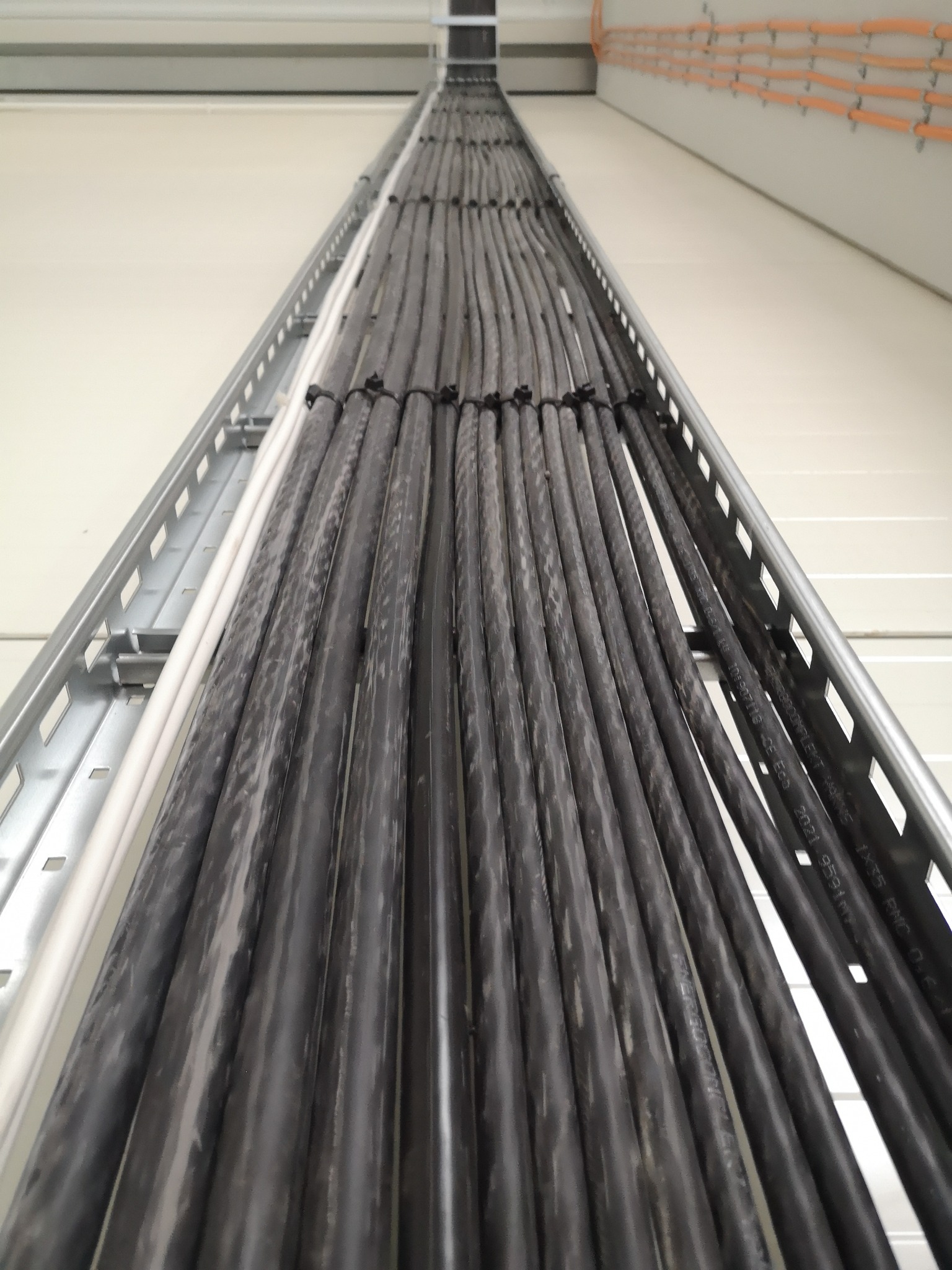 Rząd równolegle ułożonych, czarnych kabli elektrycznych, zabezpieczonych czarnymi opaskami zaciskowymi i umieszczonych w metalowej korycie kablowej, biegnącej wzdłuż jasnej ściany.