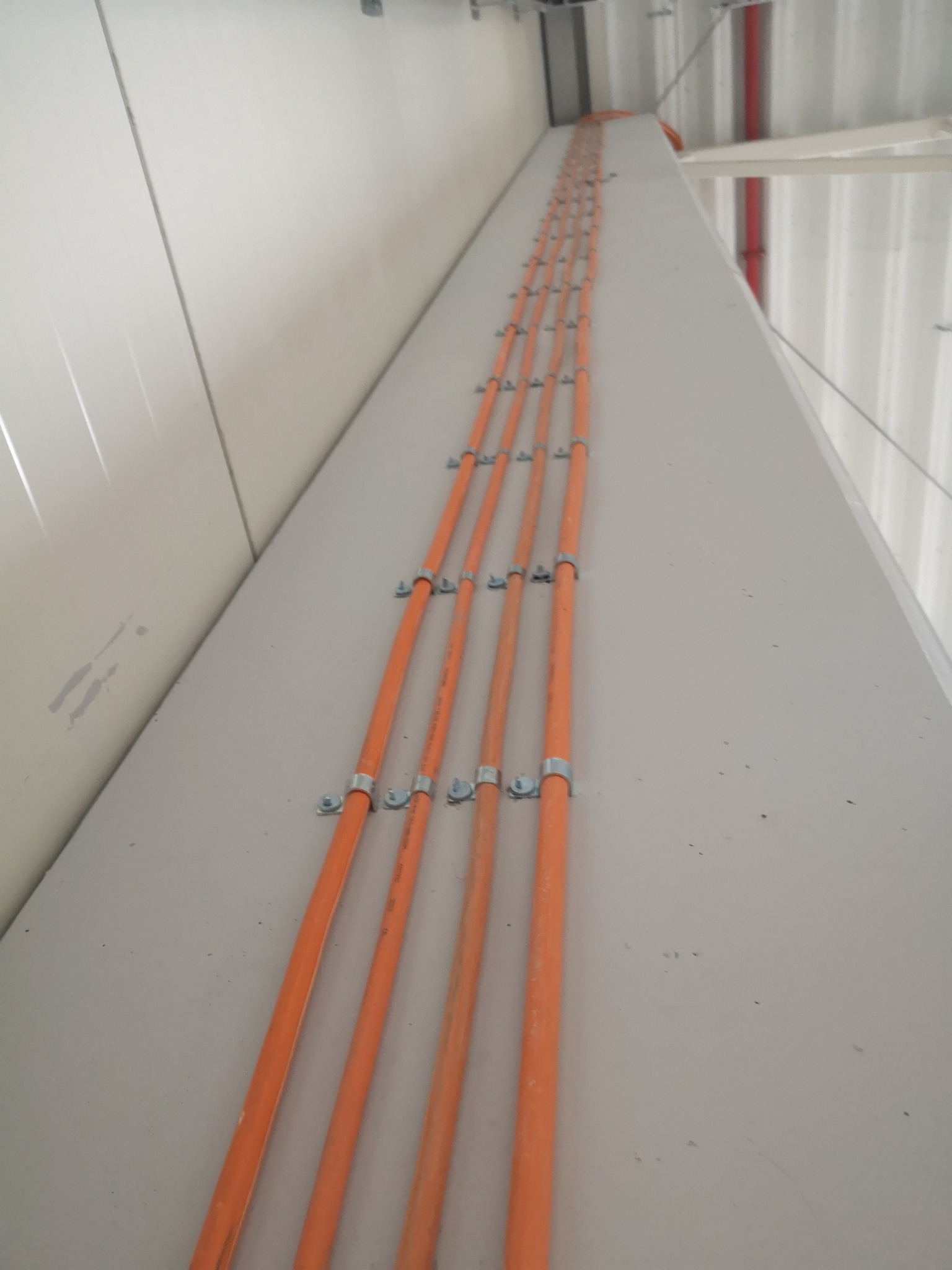 Pomarańczowe rury ułożone równolegle, przymocowane metalowymi uchwytami do jasnej ściany wzdłuż sufitu, widok z dołu.