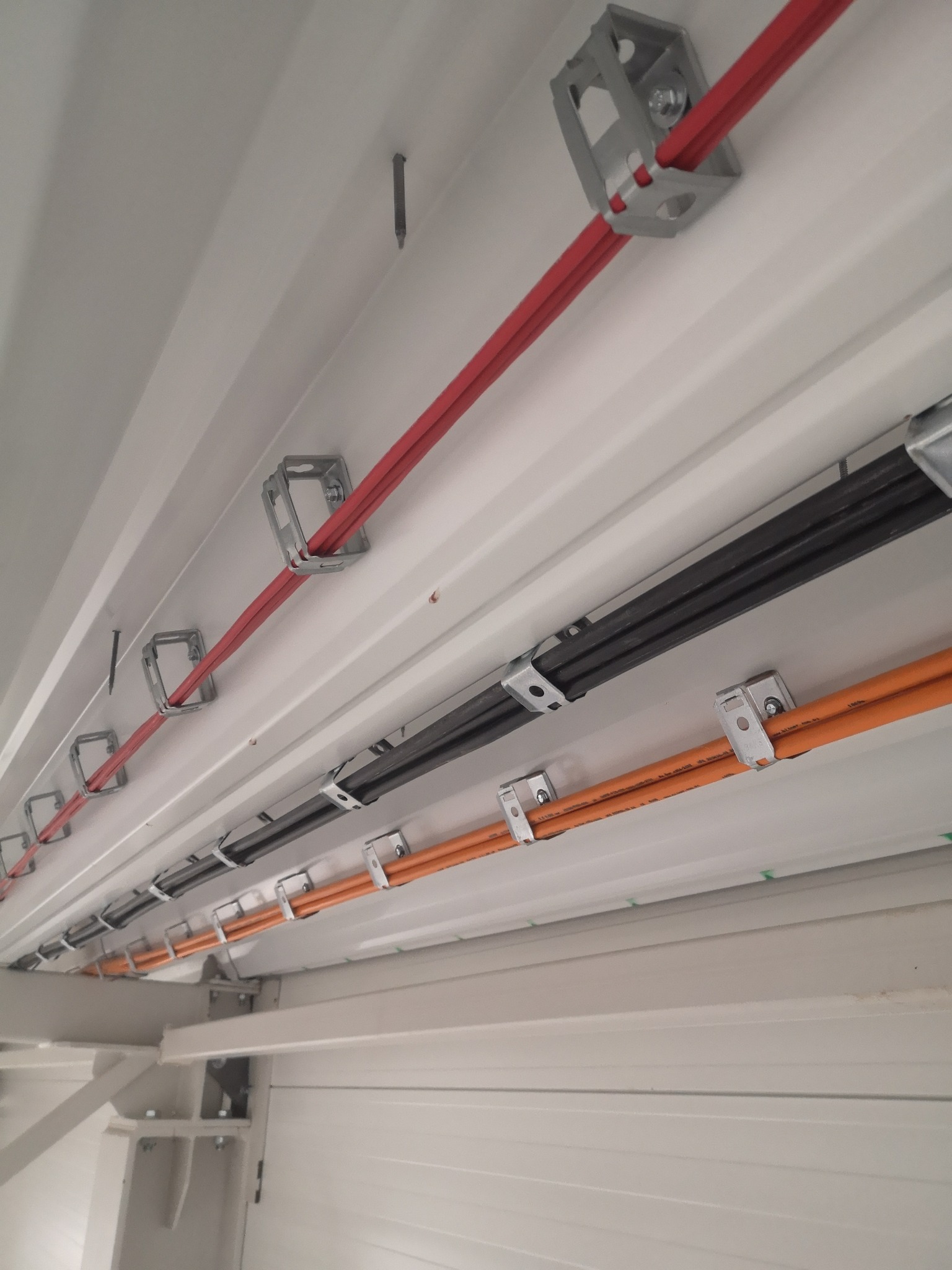 Różnokolorowe kable (czerwony, czarny, pomarańczowy) przymocowane metalowymi uchwytami do białego sufitu z blachy trapezowej w hali przemysłowej.