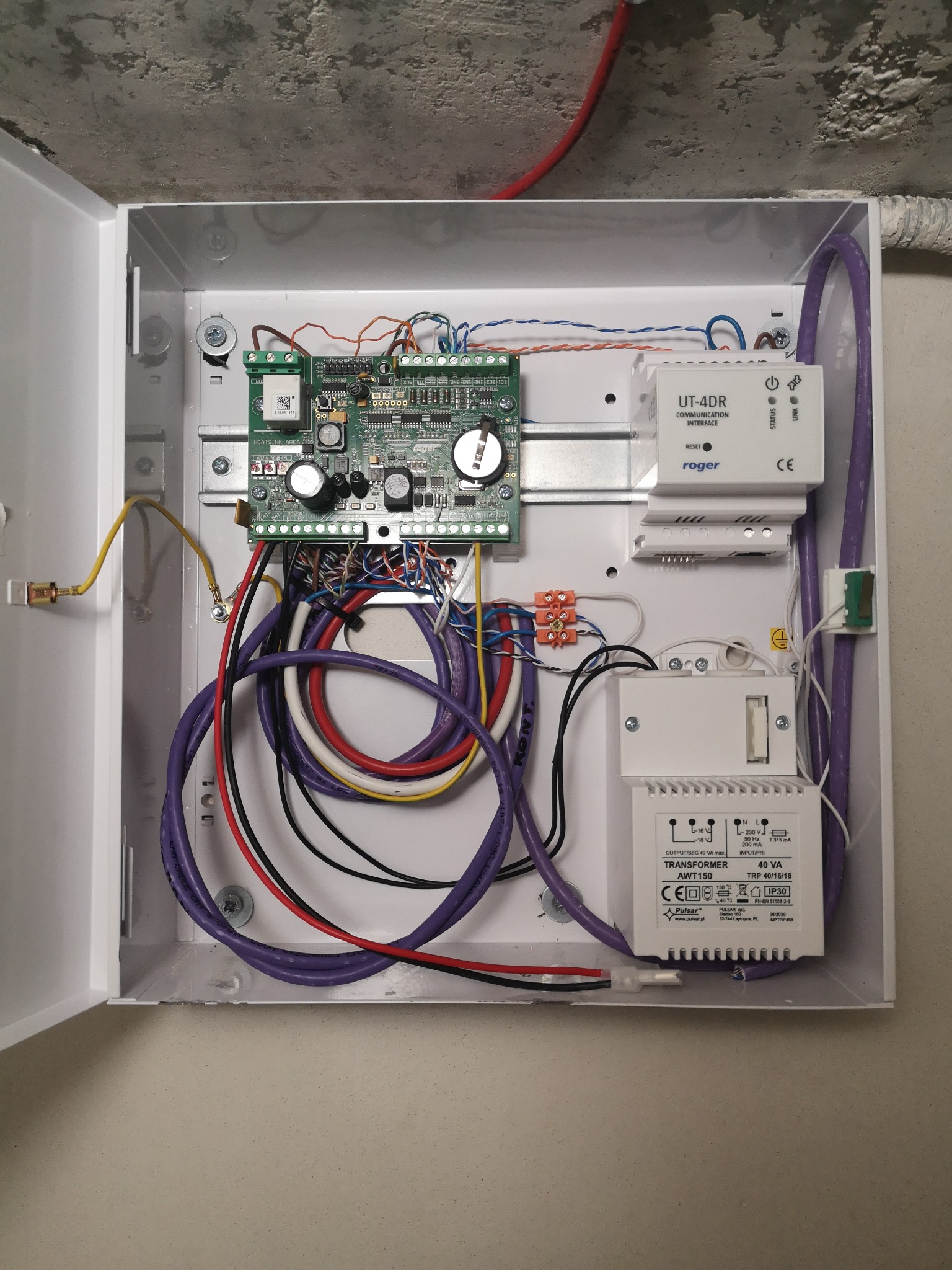 Otwarta skrzynka z elektroniką zabezpieczeń: płyta główna, transformator Pulsar AWT150, moduł komunikacyjny Roger UT-4DR oraz liczne przewody w kolorach fioletowym, białym, czarnym i czerwonym.