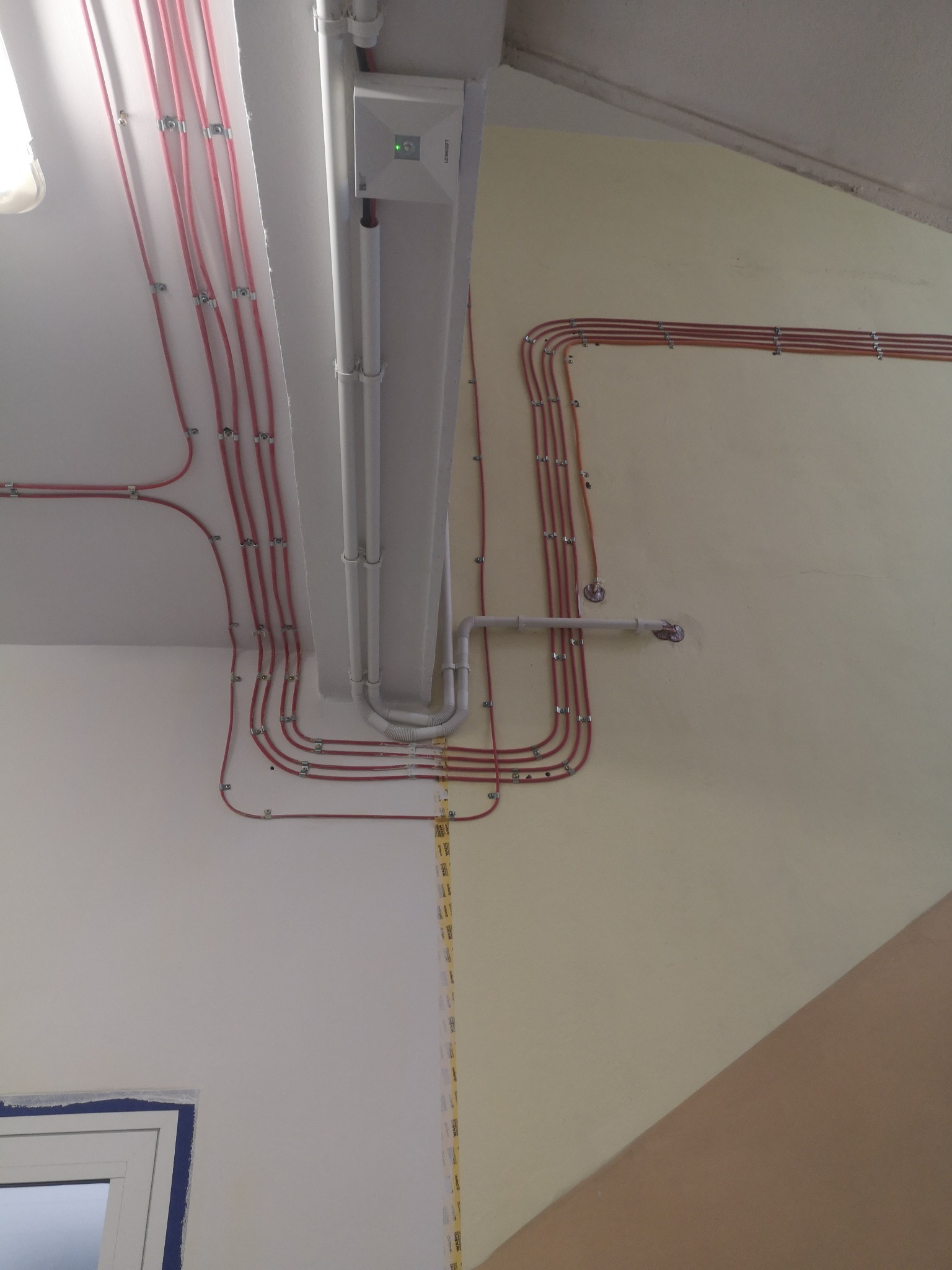 Równolegle ułożone przewody elektryczne w czerwonej izolacji, przymocowane metalowymi uchwytami do ściany i sufitu, obok białe rury i centrala alarmowa z zieloną diodą, widoczne fragment okna.