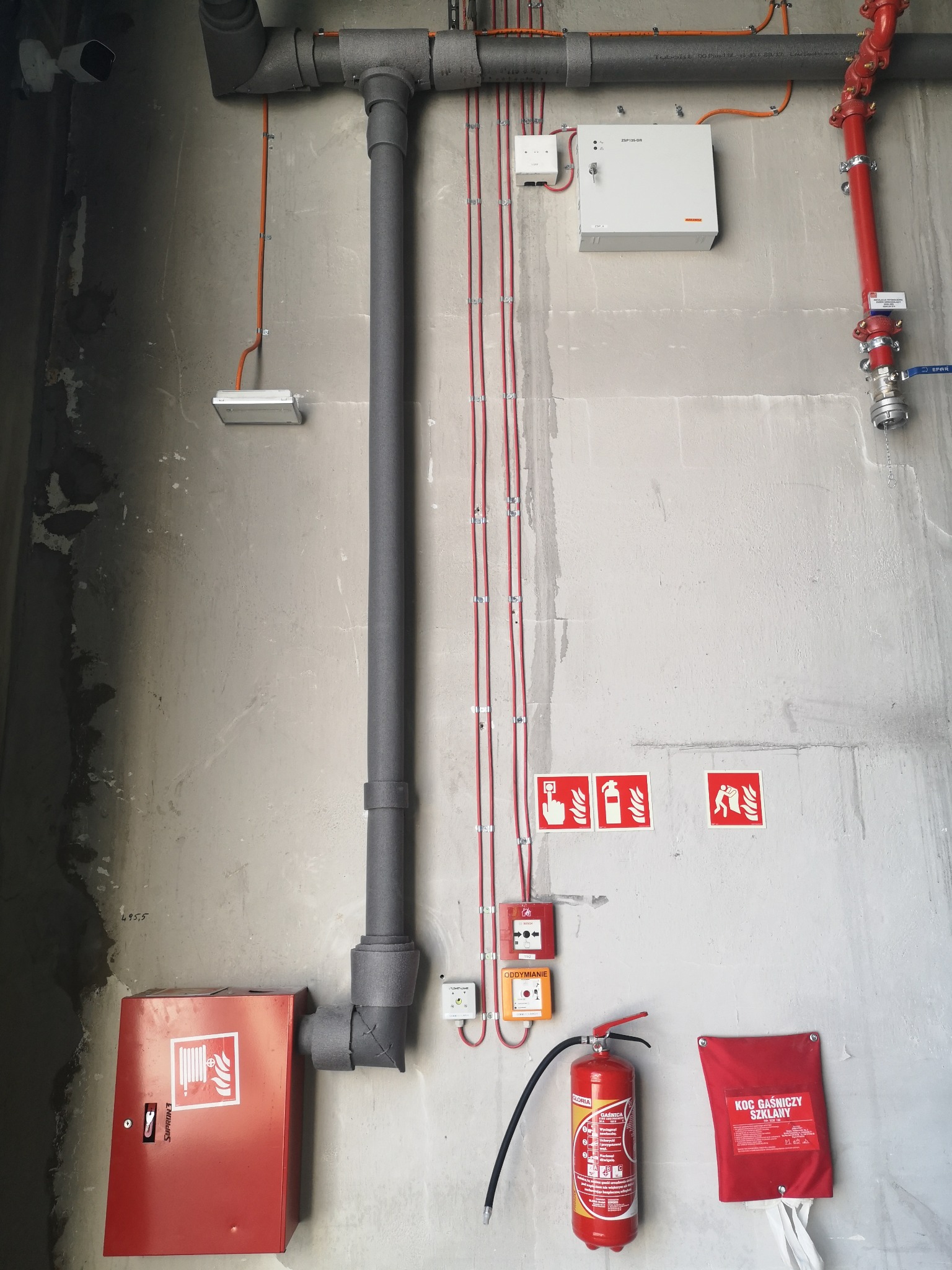 Elementy systemu przeciwpożarowego zainstalowane na surowej betonowej ścianie: rury w izolacji, czerwona instalacja hydrantowa, skrzynka na wąż strażacki, gaśnica, koc gaśniczy, czujki i przyciski...
