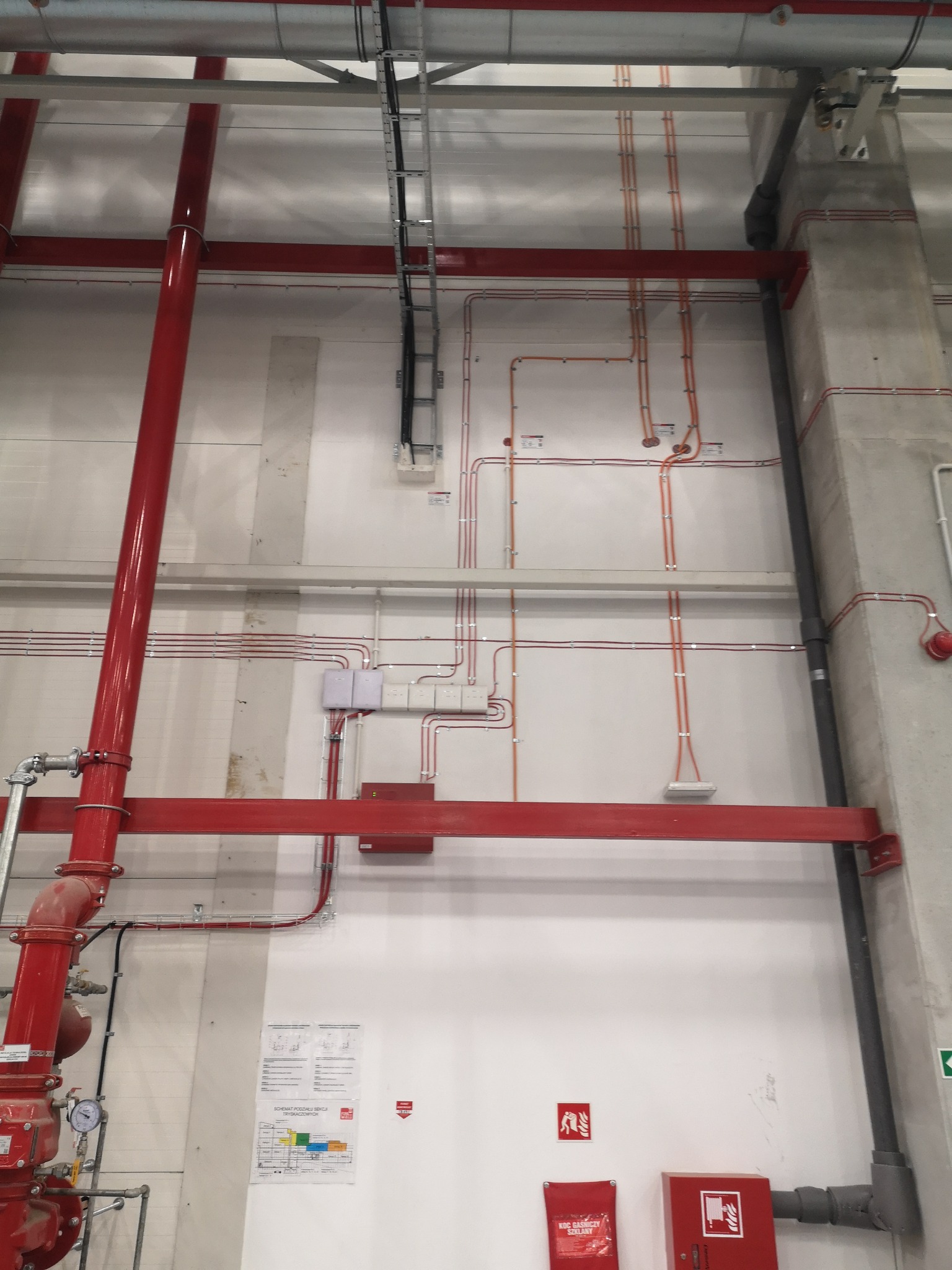 Wnętrze hali przemysłowej z widocznymi instalacjami: rurami przeciwpożarowymi w kolorze czerwonym, korytami kablowymi pod sufitem oraz okablowaniem elektrycznym prowadzonym po ścianach. Widoczne...