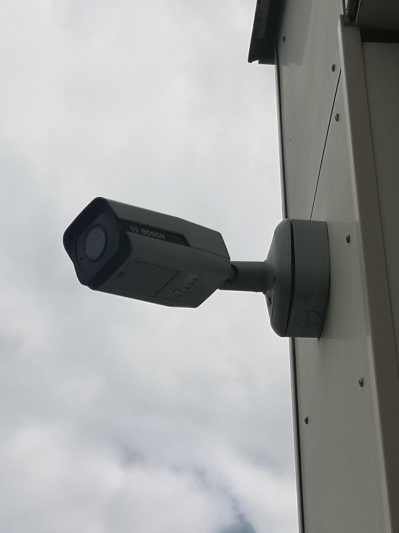 Szara kamera monitoringu marki Bosch zamontowana na jasnej ścianie budynku, widok z dołu na tle pochmurnego nieba.