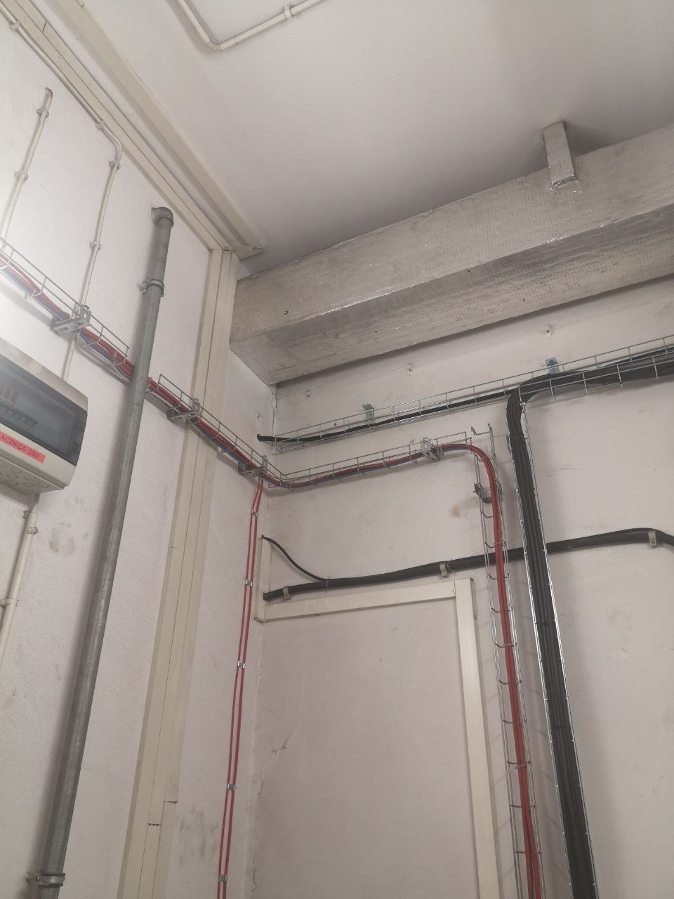 Szare pomieszczenie z widocznymi przewodami elektrycznymi ułożonymi na ścianach i suficie, z puszką rozdzielczą po lewej stronie, rury pionowe w metalowej osłonie.