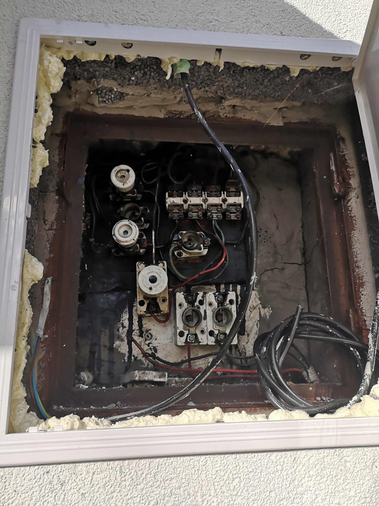 Otwarta skrzynka elektryczna z bezpiecznikami i kablami, zabezpieczona pianką montażową, widoczne ślady korozji i uszkodzeń.