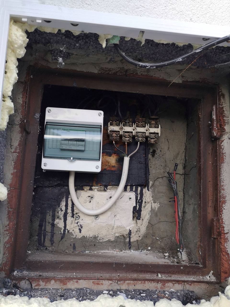 Skrzynka elektryczna z bezpiecznikami zamontowana w nieregularnym otworze w ścianie, widoczne przewody i pianka montażowa uszczelniająca krawędzie otworu.