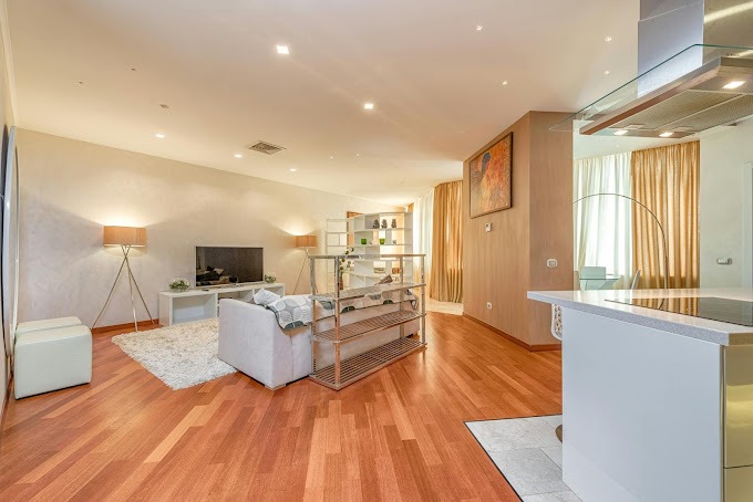 Jasne, przestronne wnętrze salonu z drewnianą podłogą, białym dywanem i sofą, widoczne lampy na trójnogach i część aneksu kuchennego z okapem.