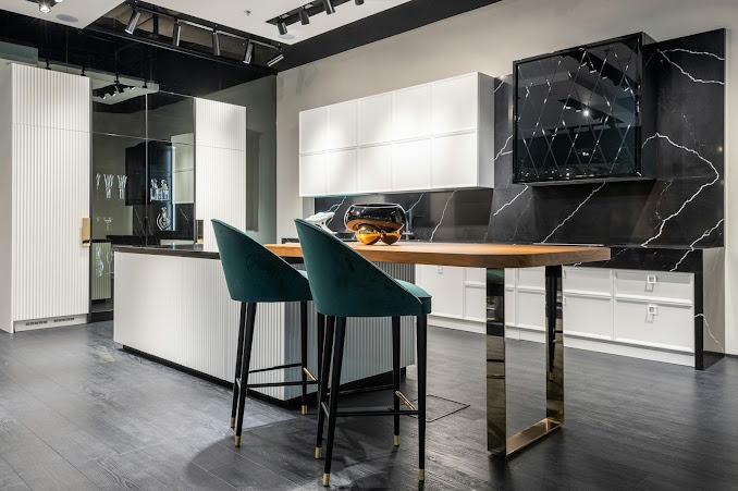 Nowoczesna kuchnia z białymi szafkami, blatem z naturalnego drewna i czarnym marmurem na ścianie, uzupełniona turkusowymi hokerami z czarnymi nogami i złotymi detalami.