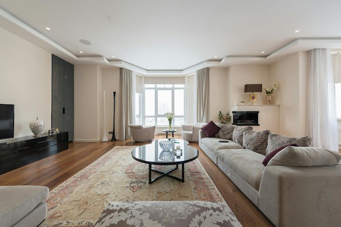 Przestronny salon z drewnianą podłogą, dywanem w orientalne wzory, szarą sofą modułową z poduszkami, okrągłym stolikiem kawowym ze szklanym blatem, zabudową meblową w ciemnym kolorze oraz dużym...