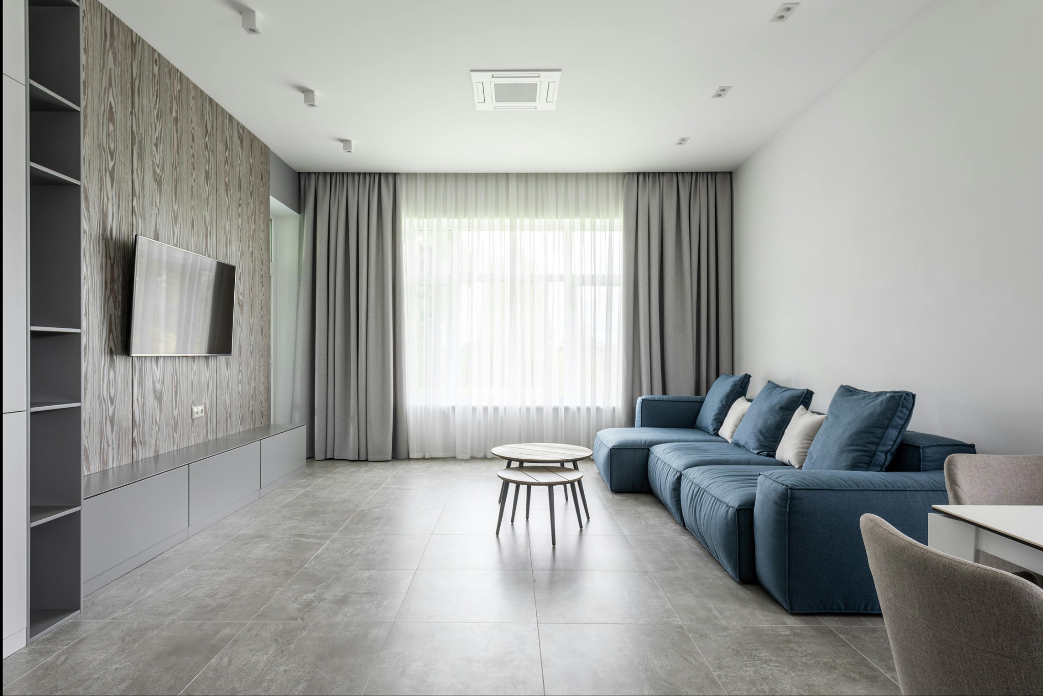 Nowoczesny salon z niebieską sofą modułową, stolikiem kawowym z dwoma blatami i ścianą z okładziną imitującą drewno z wbudowanym regałem i telewizorem.