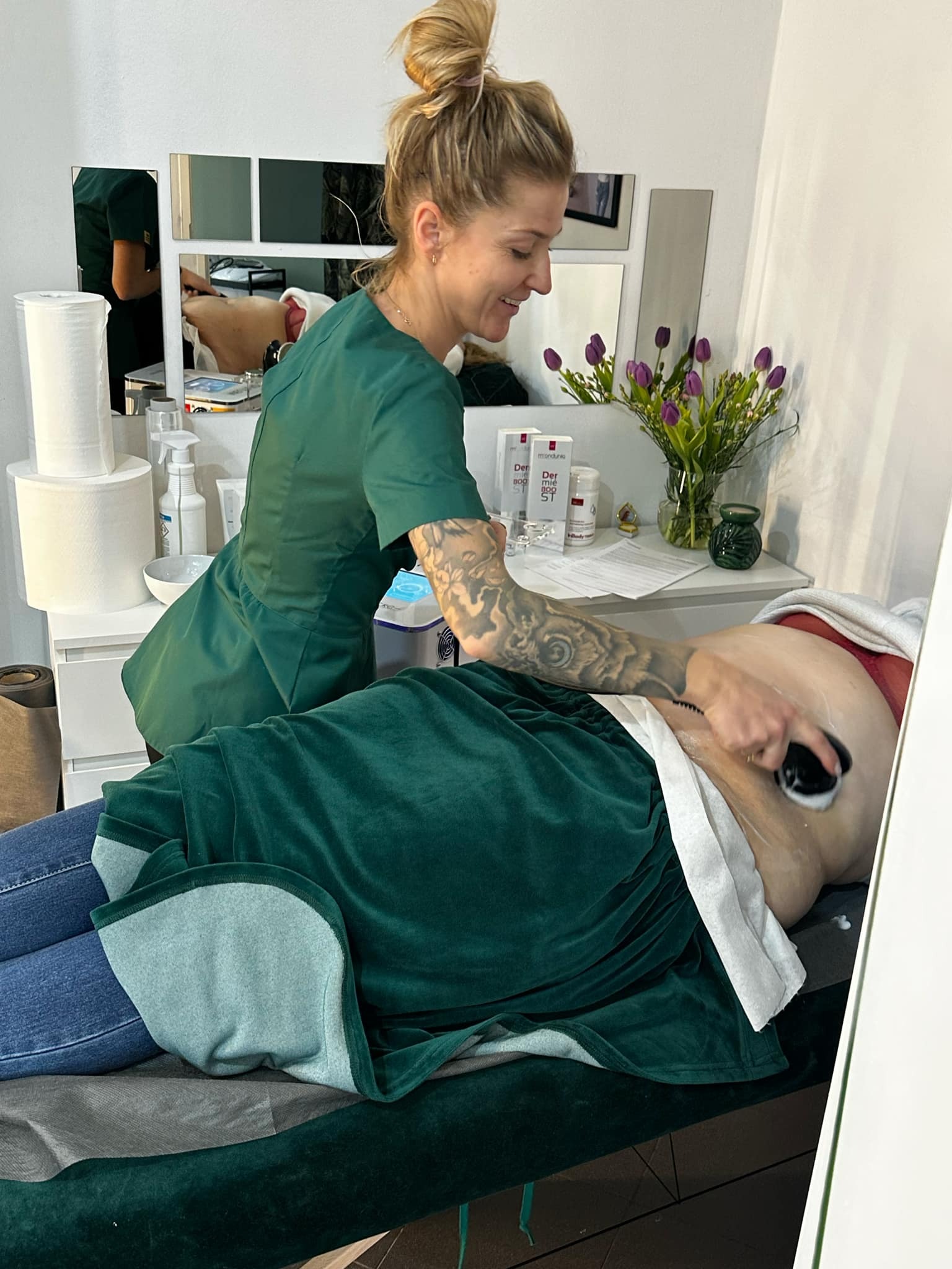 Kobieta w zielonym uniformie wykonuje masaż podciśnieniowy brzucha klientki leżącej na zielonym, welurowym leżaku; widoczne kosmetyki i bukiet tulipanów w tle.