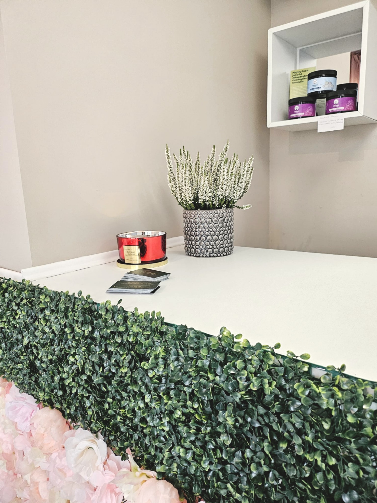 Dekoracyjny stół w salonie kosmetycznym: kwiaty, świeca zapachowa i kosmetyki na półce. Elementy wystroju tworzą relaksującą atmosferę.