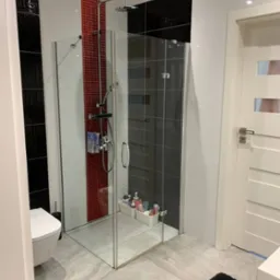 Nowoczesna kabina prysznicowa z przezroczystego szkła z chromowaną armaturą w łazience z czarno-biało-czerwoną mozaiką, biała toaleta i czarny dywanik na szarych płytkach podłogowych.