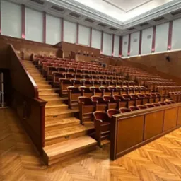 Wnętrze sali widowiskowej z rzędami drewnianych krzeseł z czerwonym obiciem, parkiet w jodełkę na podłodze, drewniane schody i jasne ściany z dekoracyjnymi elementami.