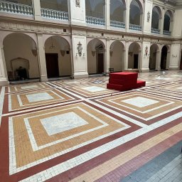 LAVIAN - Wnętrze przestronnego holu z mozaikową podłogą w geometryczne wzory, arkadowe przejścia i czerwona konstrukcja w centrum, widoczne ślady po remoncie.