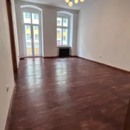 Pusty pokój z odnowioną, ciemnobrązową, lakierowaną podłogą, białymi ścianami, dwoma oknami z widokiem na budynki i czarnym grzejnikiem pod jednym z okien, oraz nowoczesnym, czarnym żyrandolem...