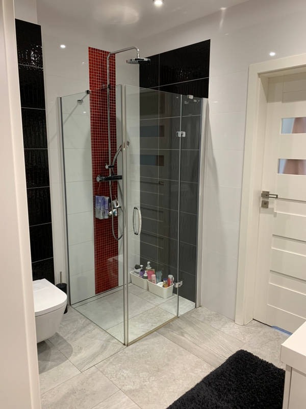 Nowoczesna kabina prysznicowa z przezroczystego szkła z chromowaną armaturą w łazience z czarno-biało-czerwoną mozaiką, biała toaleta i czarny dywanik na szarych płytkach podłogowych.