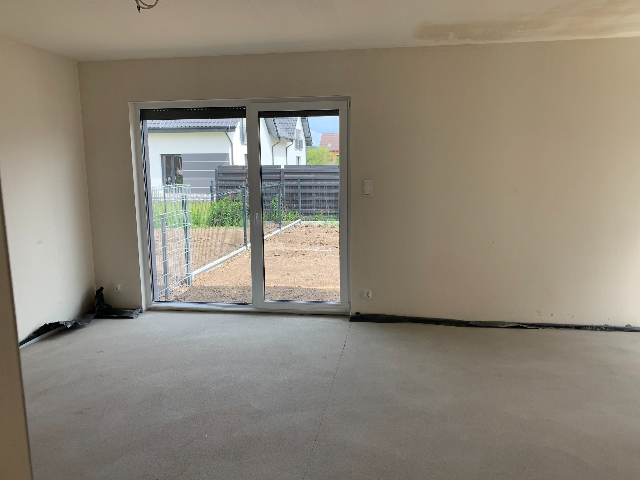 Surowe wnętrze nowego mieszkania z wylewką podłogową i zabezpieczeniem ścian folią, widok na ogród przez przesuwne drzwi balkonowe.