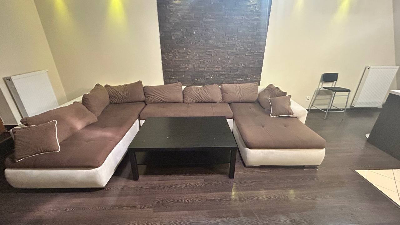 Ustawny narożnik w odcieniach brązu z niskim, czarnym stolikiem kawowym na parkiecie, ściana z dekoracyjnej cegły w tle, widoczne dwa grzejniki i krzesło.