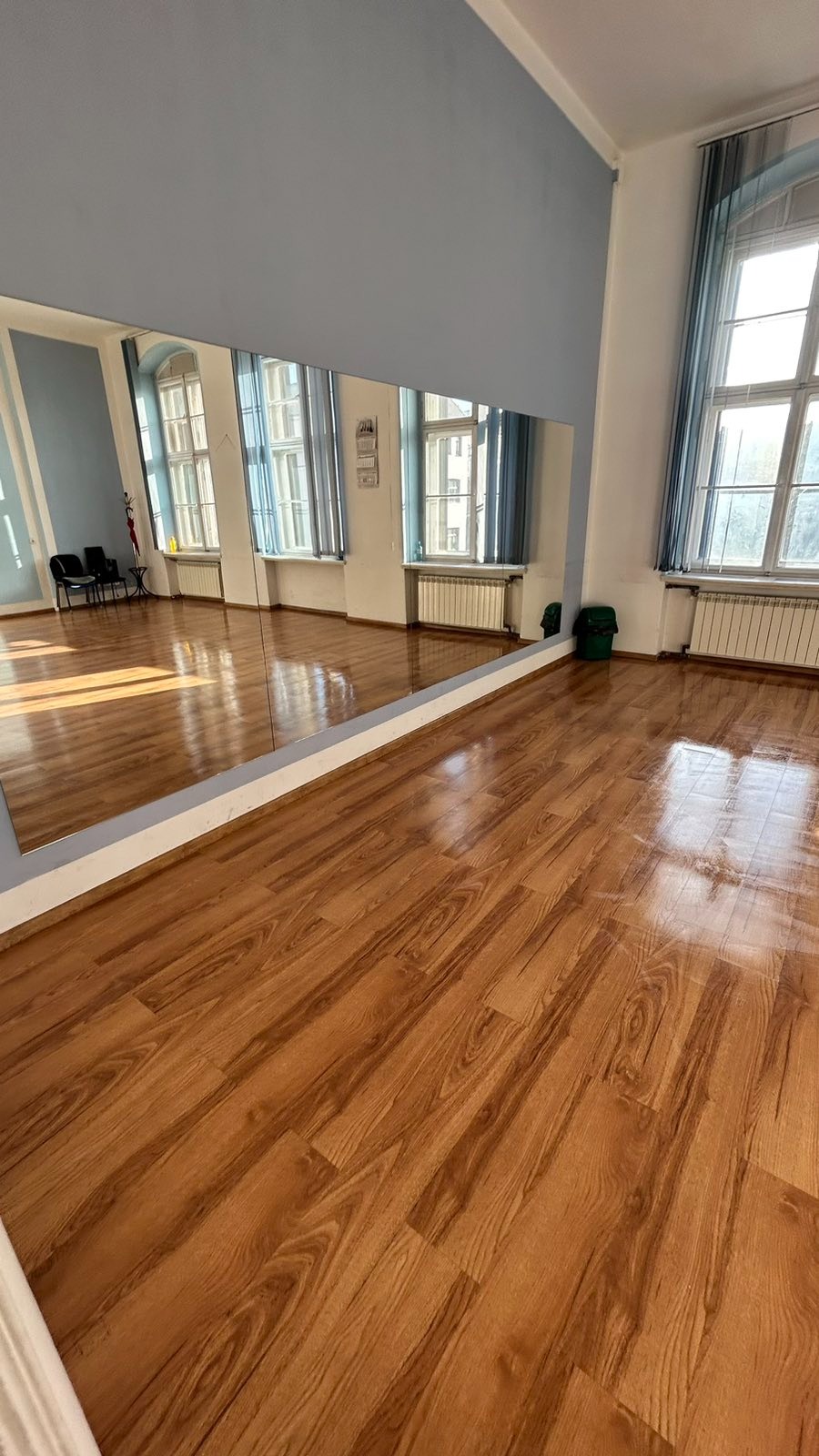 Błyszcząca, świeżo umyta podłoga z paneli w sali tanecznej z dużym lustrem na ścianie i oknami z niebieskimi zasłonami.