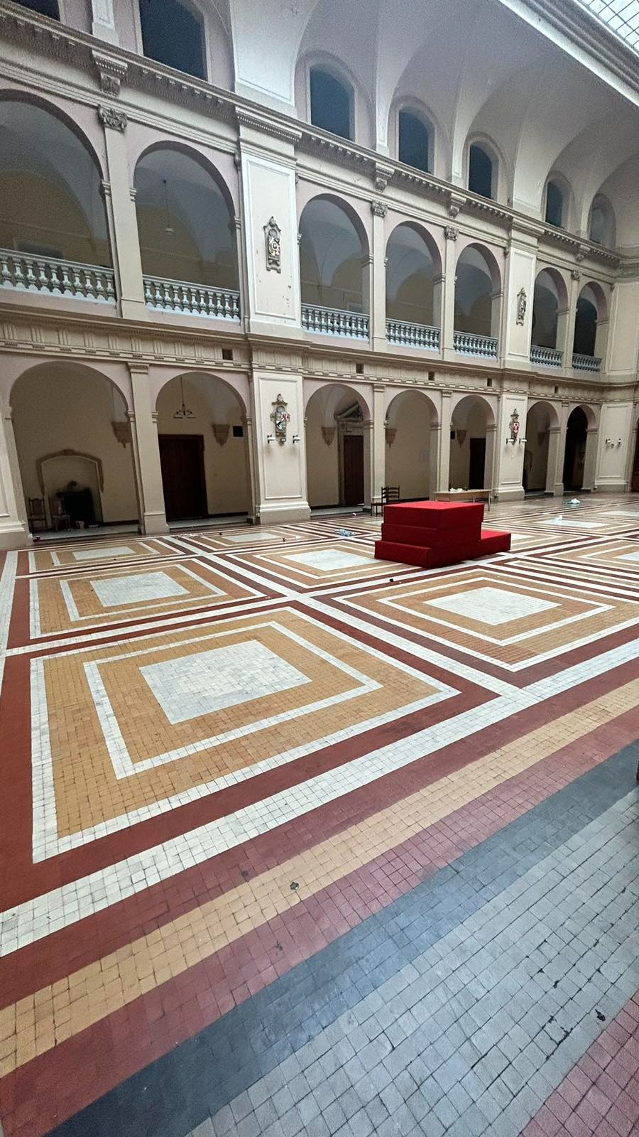 Wnętrze przestronnego holu z mozaikową podłogą w geometryczne wzory, arkadowe przejścia i czerwona konstrukcja w centrum, widoczne ślady po remoncie.