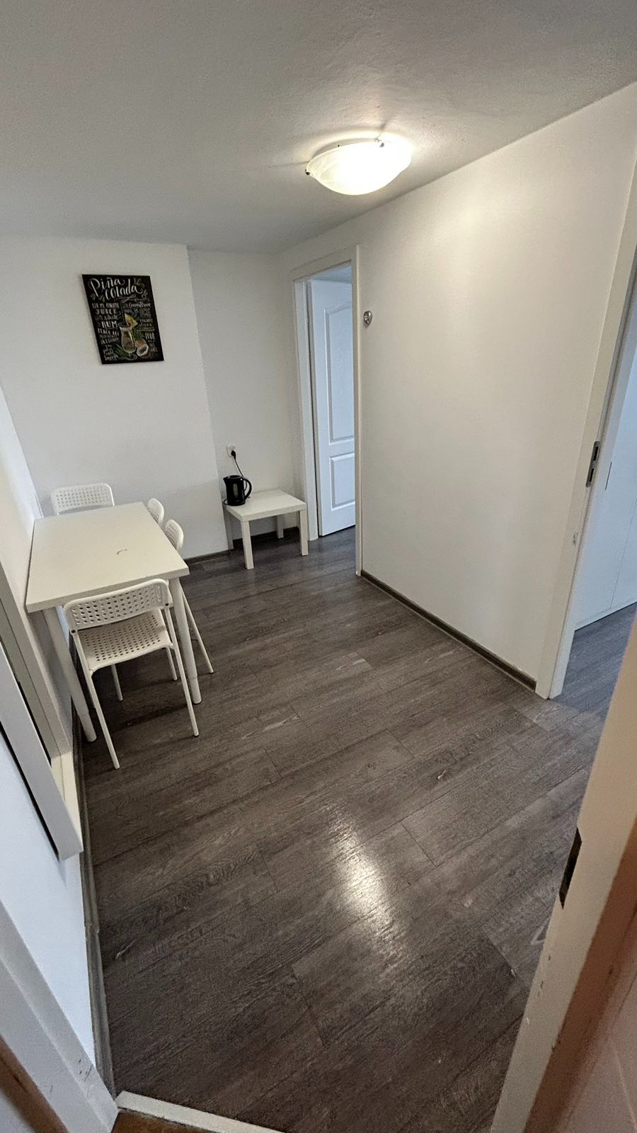 Wnętrze pokoju z białym stołem, krzesłami, ciemną podłogą imitującą drewno i plakatem z napojem Pina Colada na ścianie.