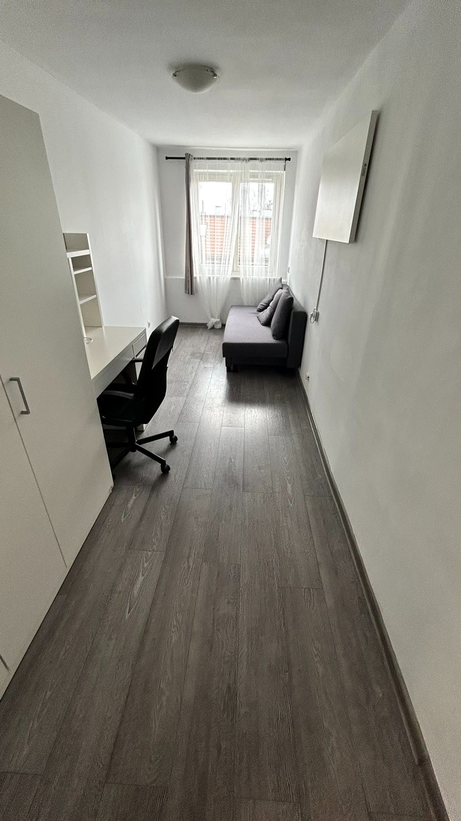 Wąski pokój z biurkiem, krzesłem obrotowym i rozkładaną sofą pod oknem z białymi firankami, oświetlony lampą sufitową i panelem ściennym.