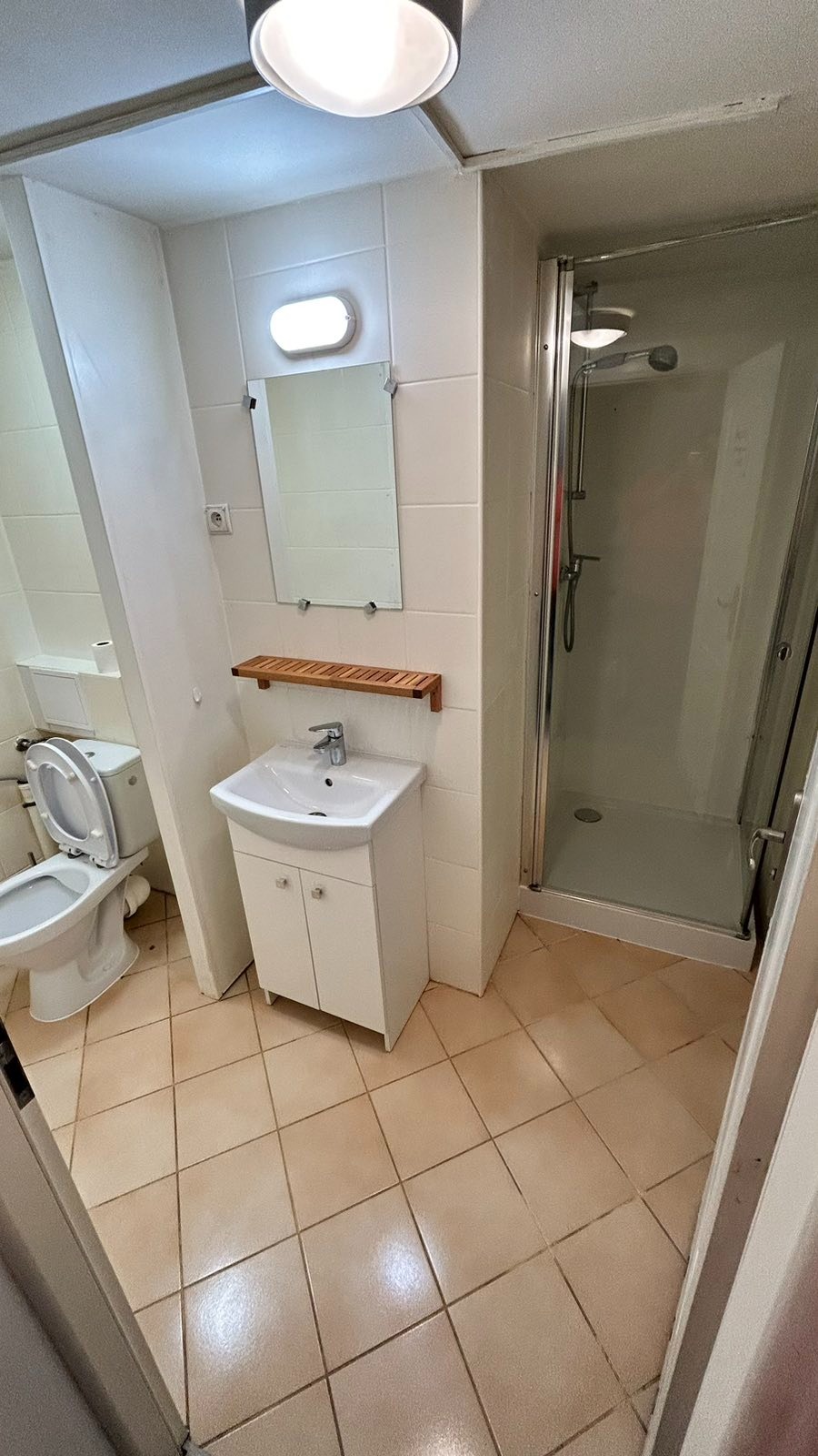 Wnętrze małej łazienki z kabiną prysznicową, umywalką z szafką i toaletą, wykończonej w jasnych kolorach z beżową terakotą na podłodze.