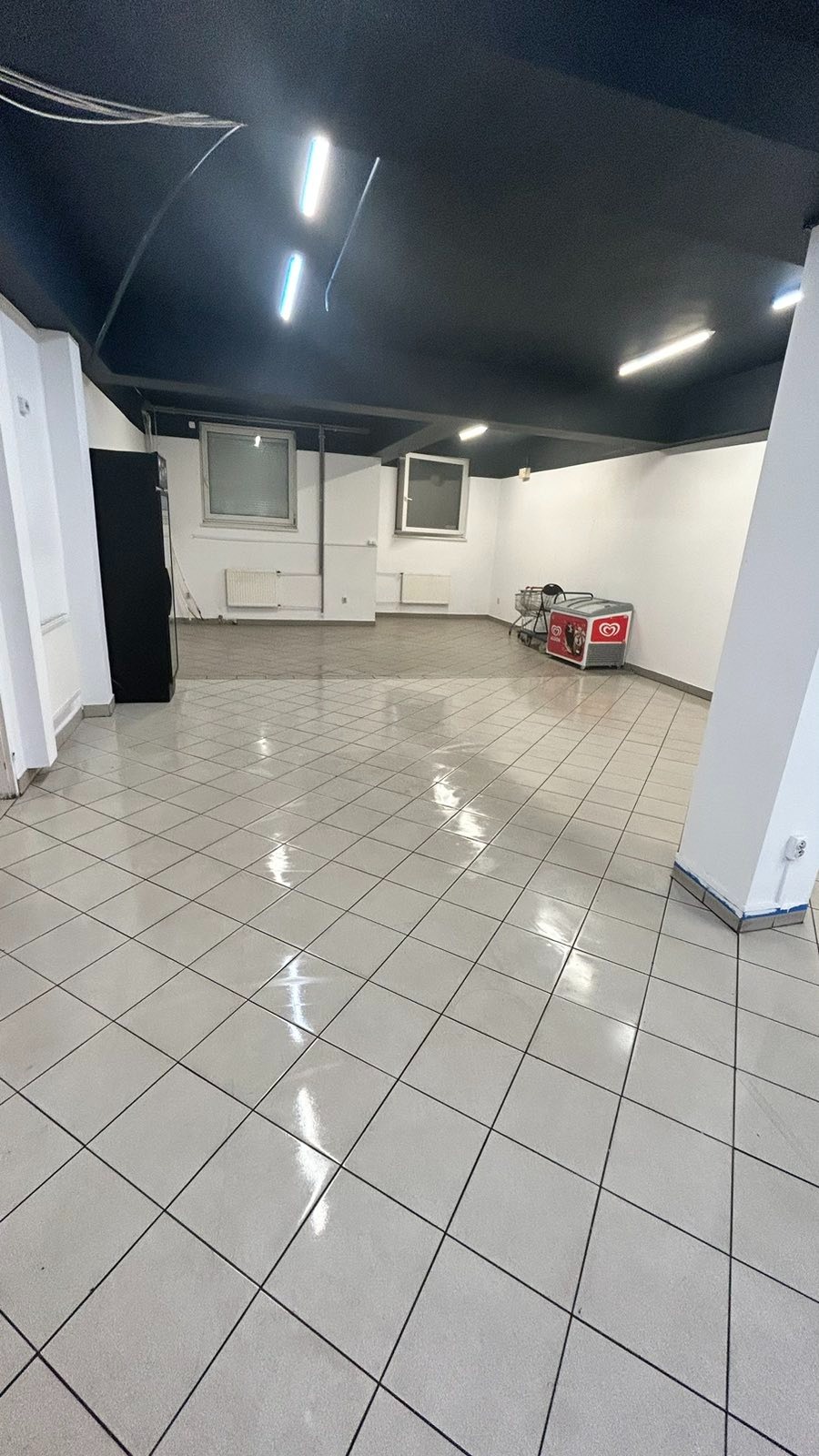 Pusta przestrzeń biurowa z białą podłogą z płytek, czarnym sufitem, metalową szafką, wózkiem sklepowym i lodówką do lodów.